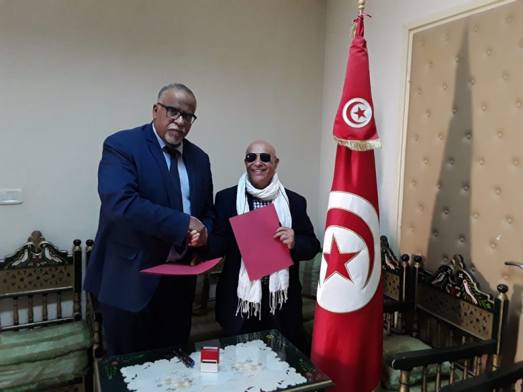 Vereinsgründer Dr. Mustapha Ouertani schüttelt die Hand eines Vertreters der örtlichen Schulbehörde, welcher sich auf der linken Seite des Bildes befindet. Beide Männer haben dünne rote Ordner in der Hand. Auf der rechten Seite von Dr. Ouertani steht eine Menschengroße Tunesien-Flagge. 