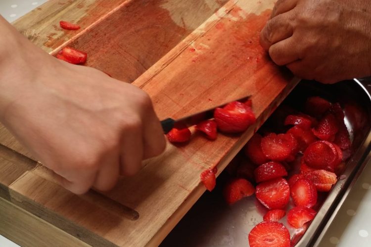 Von einem Holzbrett werden geschnittene Erdbeeren in eine Schüssel geschoben.