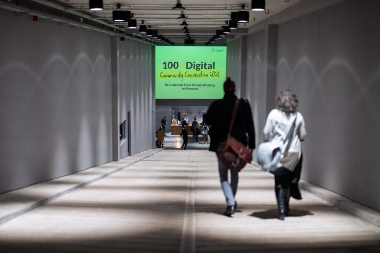 Zwei Menschen laufen einen langen Gang hinunter. An einer Wand am Ende des Ganges steht die Beschriftung 100xDigital Community Convention 2023 – Das Netzwerk-Event für Digitalisierung im Ehrenamt.