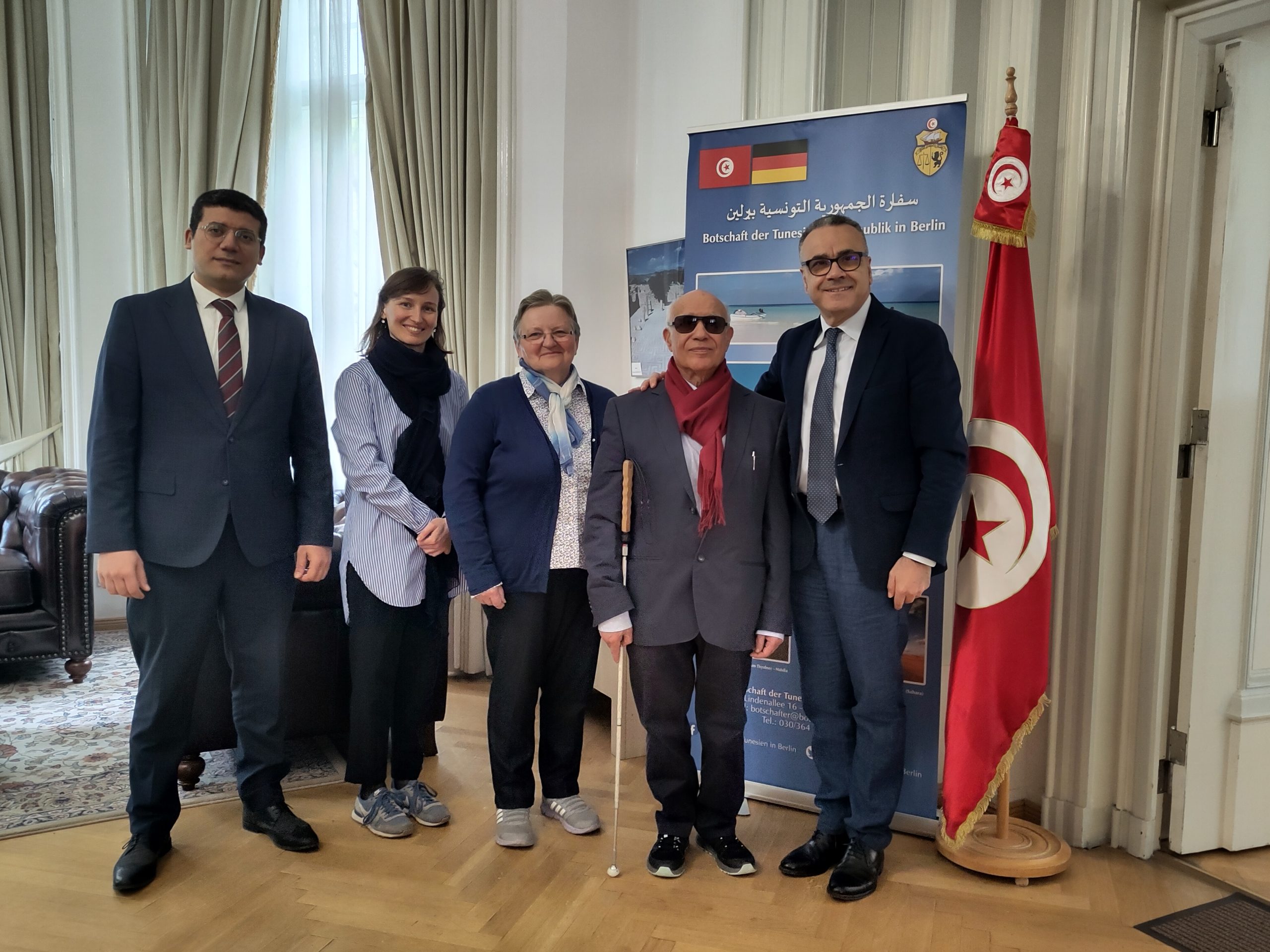 Gruppenfoto des Vereinsgründers Dr. Mustapha Ouertani und der Vorstandsvorsitzenden Frau Smyrek-Ouertani mit dem tunesischen Botschafter und dem Kulturattachée in Deutschland vor einer großen tunesischen Flagge.